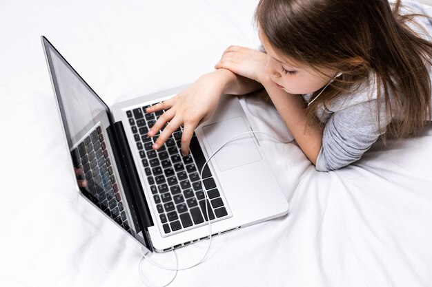 Niña linda que escribe con los dedos en las teclas de la computadora portátil en la sábana blanca en la cama en la vista superior de casa