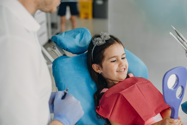 Niña linda que mira en el espejo mientras está sentado en una silla de estomatología después de realizar una cirugía de dientes.