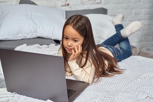 Niña linda que se siente divertida mientras ve dibujos animados en una computadora portátil sentada en la cama