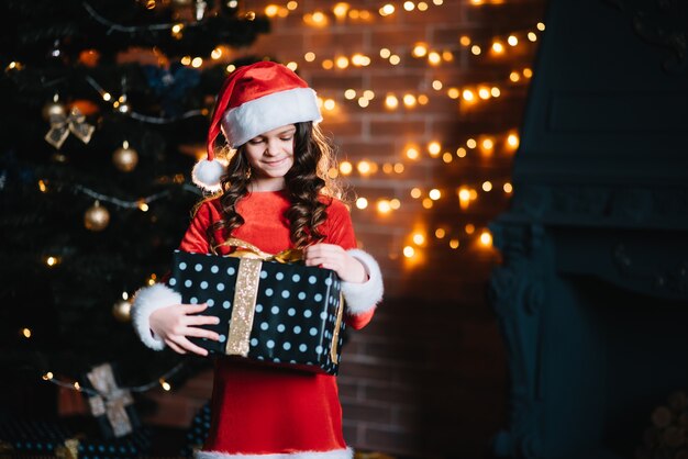 Niña linda que sostiene un regalo cerca del árbol de Navidad en un traje rojo de santa