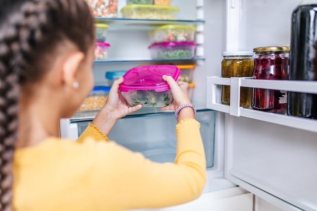 Foto niña linda que toma el recipiente con brócoli congelado del refrigerador.