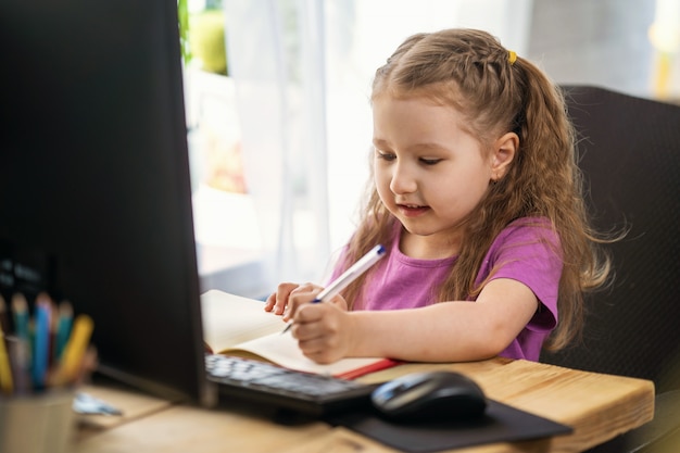 Una niña linda que usa una computadora para aprendizaje electrónico remoto, escribe en un cuaderno