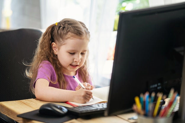 Una niña linda que usa una computadora para aprendizaje electrónico remoto, escribe en un cuaderno