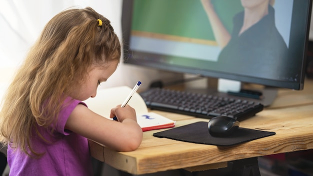 Una niña linda que usa una computadora para aprendizaje electrónico remoto, escribe en un cuaderno