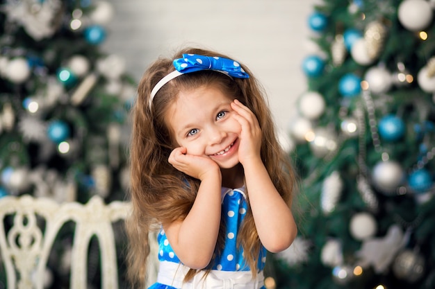 Niña linda con un regalo de Navidad, con un vestido azul se sienta junto a los árboles de Navidad decorados. Año Nuevo, vacaciones familiares de Navidad