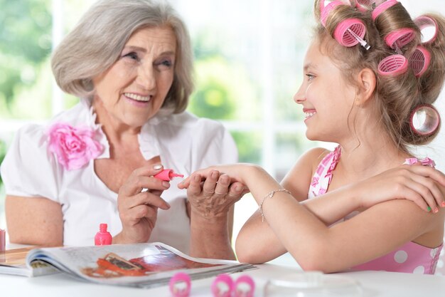 Foto niña linda en rizadores de pelo con la abuela con revista y esmalte de uñas