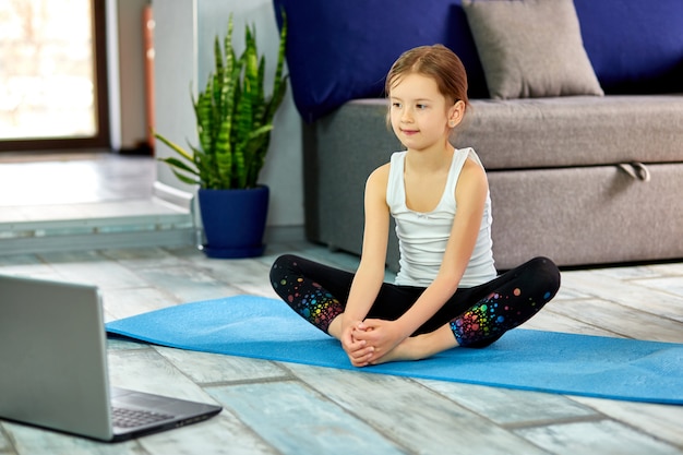 Niña linda en ropa deportiva sobre estera azul practicando gimnasia en casa