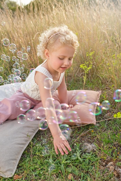 Niña linda con ropa elegante jugando con pompas de jabón mientras se sienta en almohadas acogedoras en un claro de hierba en el campo