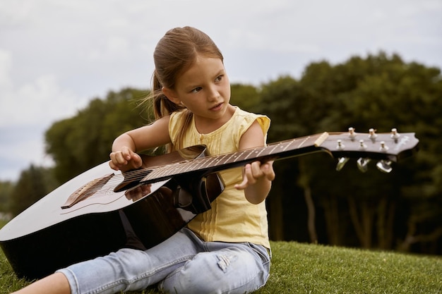 Niña linda con ropa informal sentada en un césped verde en el parque y tocando la guitarra gastando