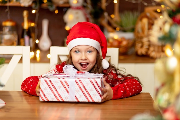 Niña linda en ropa de invierno sombrero de Santa Claus y suéter rojo se regocija con el regalo y se ríe de felicidad en la cocina oscura de la casa, el concepto de año nuevo y Navidad