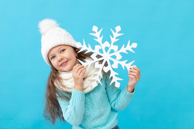 Niña linda con ropa de invierno sombrero y suéter con un gran copo de nieve en las manos sobre un fondo azul aislado sonriendo un lugar o espacio para texto