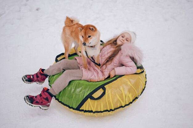 Niña linda en ropa rosa cálida divirtiéndose con el perro rojo shiba inu paseos en tubo de nieve inflable