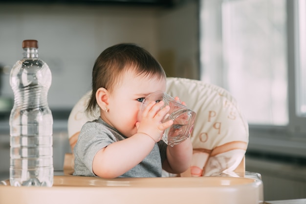 Niña linda sentada en una silla de bebé en la cocina y agua potable