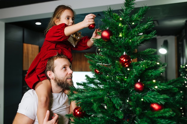 Niña linda sentada sobre los hombros de su papá decorando un árbol de navidad con bolas rojas brillantes. Papá mirando lo que está haciendo.