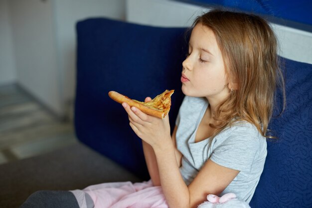 Niña linda sentada en el sofá y comiendo un trozo de pizza italiana en casa