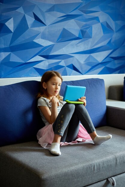 Niña linda sentada en el sofá, niño adicto a la tecnología, disfrutando de jugar juegos en línea en una tableta digital, usando aplicaciones