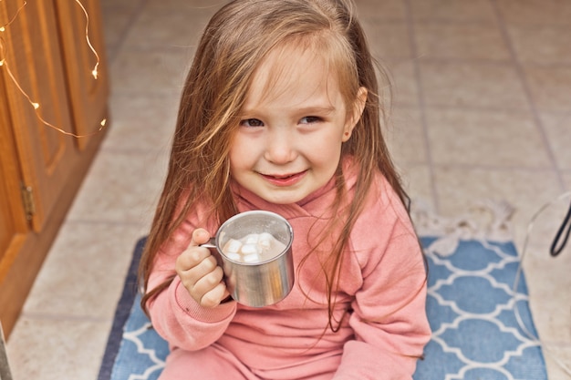 La niña linda se sienta en los escalones de un remolque de viaje y bebe chocolate caliente con malvaviscos