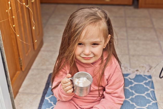 La niña linda se sienta en los escalones de un remolque de viaje y bebe chocolate caliente con malvaviscos