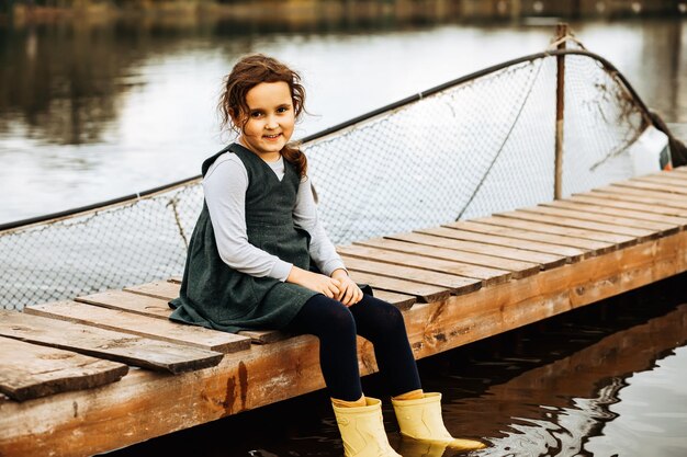 Una niña linda se sienta en un muelle de madera junto al lago, río o estanque Temporada de otoño