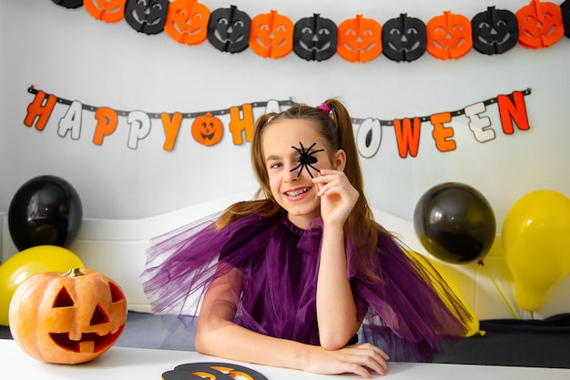 Niña linda con sombrero de bruja sentada detrás de la mesa en la habitación decorada con tema de Halloween Covid19