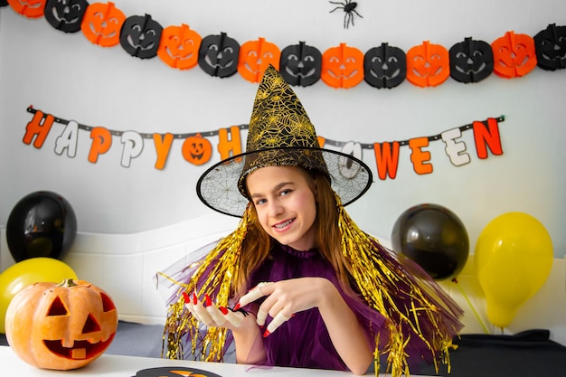 Niña linda con sombrero de bruja sentada detrás de la mesa en la habitación decorada con tema de Halloween Covid19