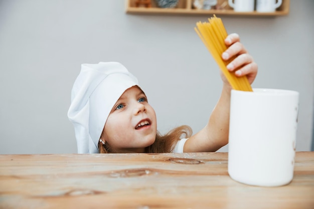 Niña linda con sombrero de chef blanco sosteniendo espagueti y lo pone en una taza grande