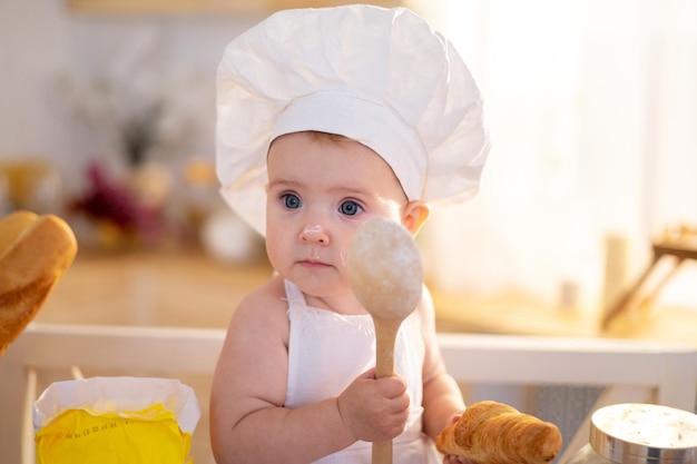 Una niña linda con un sombrero de chef y un delantal está sentada en la mesa de la cocina en casa con un rodillo de harina, hogazas de pan, un poco de comida casera para cocinar