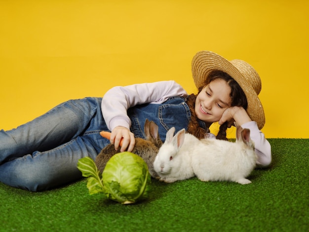 Niña linda con sombrero de paja acostada jugando con los conejitos en el fondo amarillo del estudio