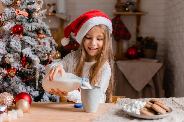 Foto una niña linda con sombrero de papá noel y pijama bebiendo leche en la cocina para una maqueta de navidad