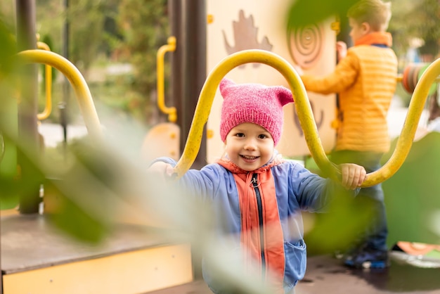 Niña linda con sombrero rosa mirando a la cámara divirtiéndose jugando en un parque infantil moderno en el parque de la ciudad