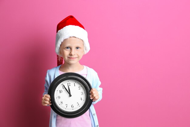 Niña linda con sombrero de Santa con reloj sobre fondo de color. Concepto de cuenta regresiva de Navidad