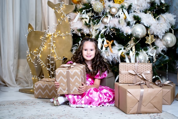 Niña linda sonriente cerca de los regalos y del árbol de navidad cercanos.