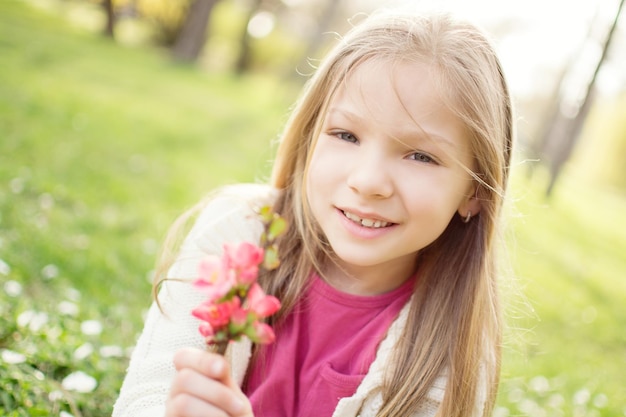 Niña linda sonriente en el prado en primavera sosteniendo flores y mirando a la cámara.