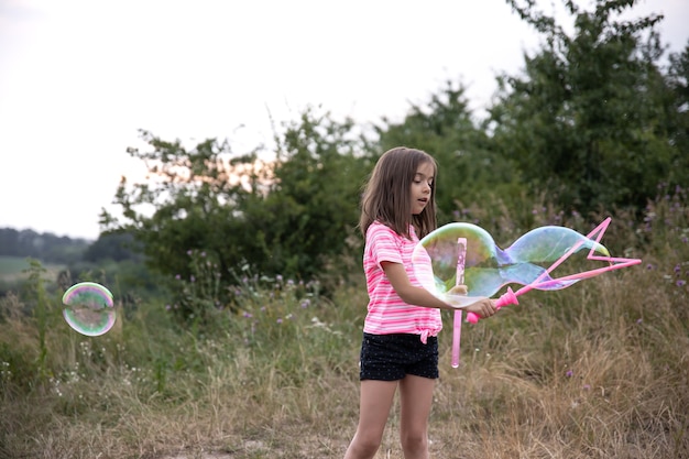 Una niña linda sopla pompas de jabón en el verano en un campo, actividades de verano al aire libre.