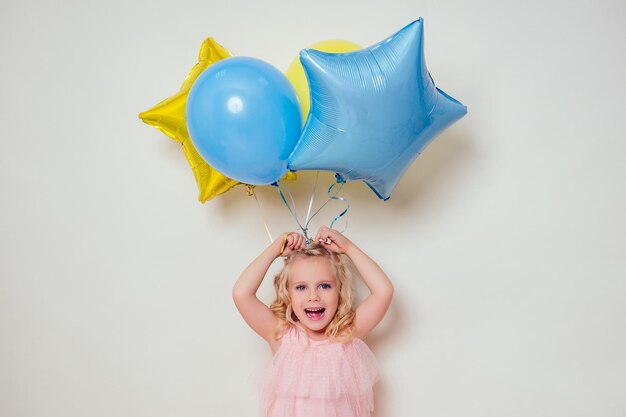 Niña linda sosteniendo globos de helio azul, amarillo y dorado sobre fondo blanco en el estudio