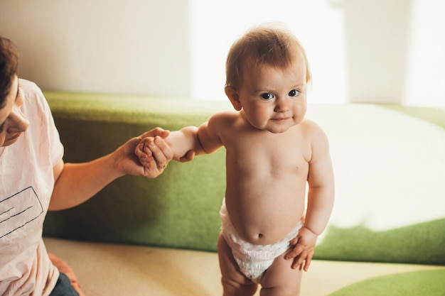 Niña linda sosteniendo la mano de mamá y mirando a la cámara mientras está de pie en el concepto de estilo de vida de la gente del sofá