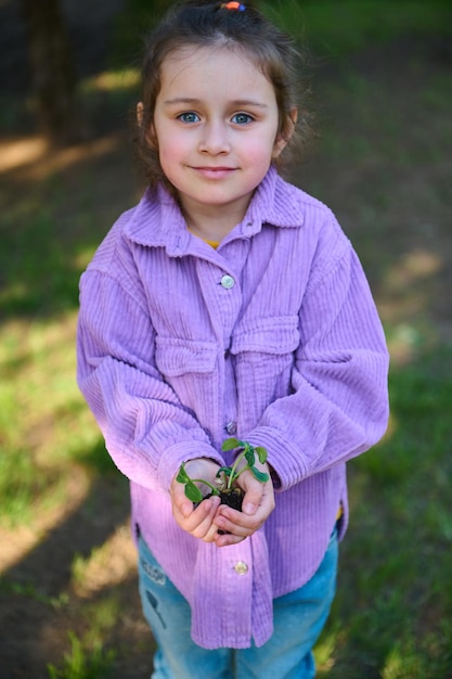 Una niña linda sostiene el suelo con brotes en las manos abiertas mira la cámara mientras está de pie en un jardín de vegetales Protección ambiental y educación cultivando vegetales orgánicos Concepto del Día de la Tierra