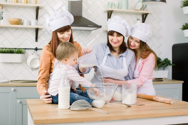 Niña linda y su hermosa mamá, tía y abuela en delantales y sombreros divirtiéndose mientras vierte leche en harina y amasando la masa en la cocina moderna en Sweet Home. Mujeres horneando en la cocina