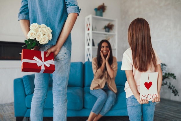 Una niña linda y su padre esconden regalos a sus espaldas mientras planean sorprender a una mujer sentada en el sofá