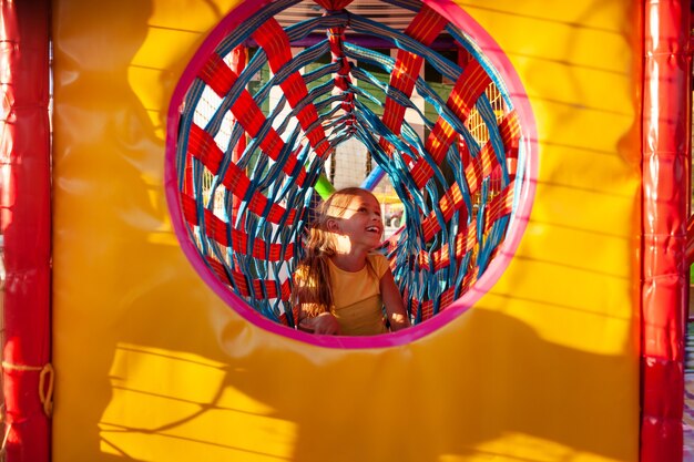 Foto niña linda sube a través de un túnel largo y brillante en un patio de recreo con coloridos equipos blandos disfrutando de las vacaciones de verano