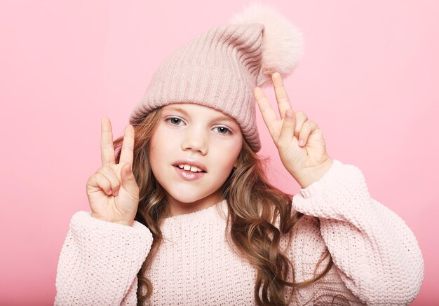 Niña linda en suéter rosa y sombrero de invierno que muestra el signo de la victoria