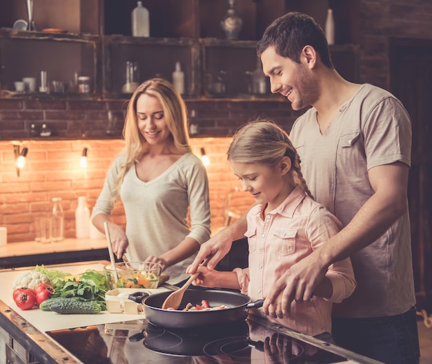 Niña linda y sus hermosos padres están sonriendo mientras cocinan en la cocina de casa