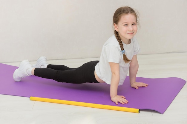 Una niña linda se para en un tablón sobre una alfombra, un palo de gimnasia se encuentra cerca, el niño sonríe