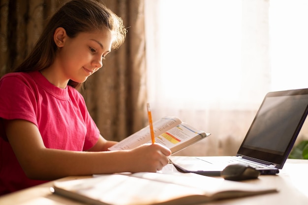 Niña linda trabajando en una computadora y escribiendo tareas en su cuaderno