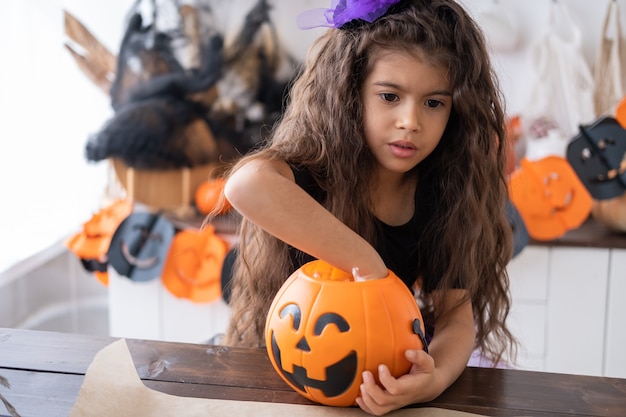 Niña linda en traje de bruja sosteniendo calabaza jack con caramelos, divirtiéndose en la cocina, celebrando Halloween.