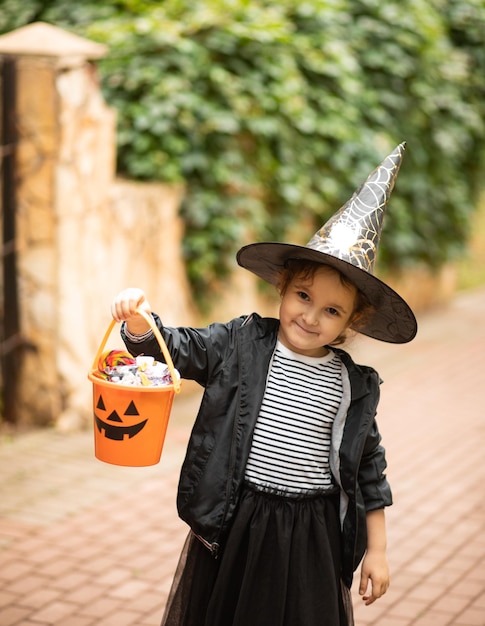 Niña linda en traje de bruja sosteniendo un cubo de calabaza jackolantern con dulces y dulces Niño truco o trato en las vacaciones de Halloween