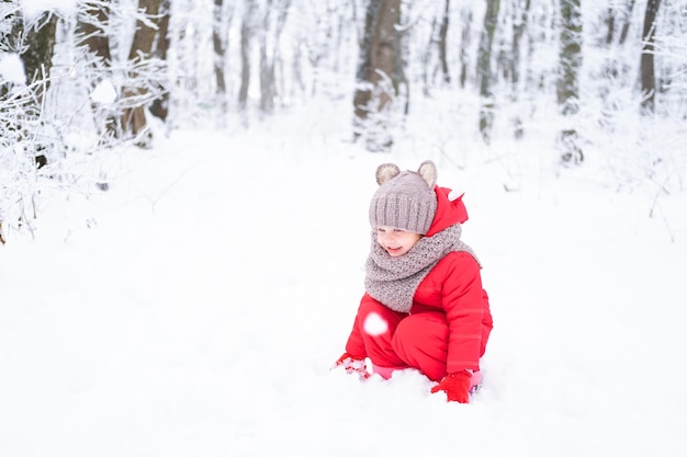 Niña linda en traje de nieve rosa juega con nieve en el bosque de invierno