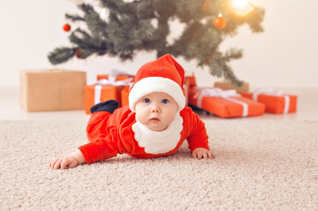 Niña linda con traje de santa claus arrastrándose por el suelo sobre el árbol de Navidad. Temporada de vacaciones.