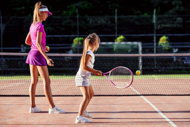 Niña linda en uniforme blanco golpeando la pelota debajo de la red durante el entrenamiento de tenis con el entrenador. Entrenador femenino atractivo que hace ejercicios con el pequeño jugador de tenis en la cancha al aire libre al atardecer.