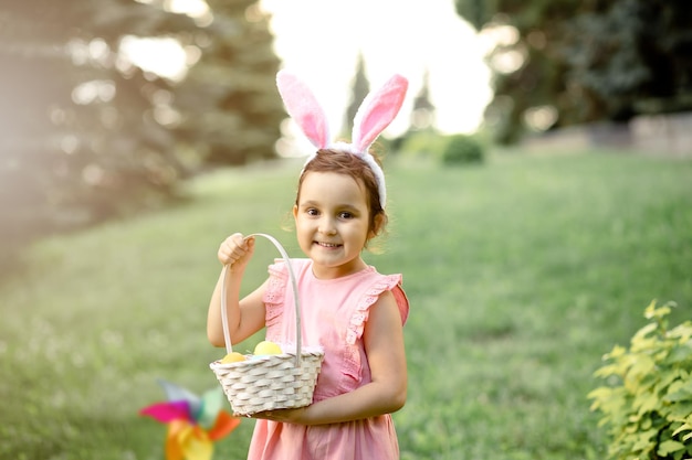 Una niña linda usa orejas de conejo sosteniendo una canasta con huevos pintados de colores en la búsqueda de huevos de Pascua en el parque